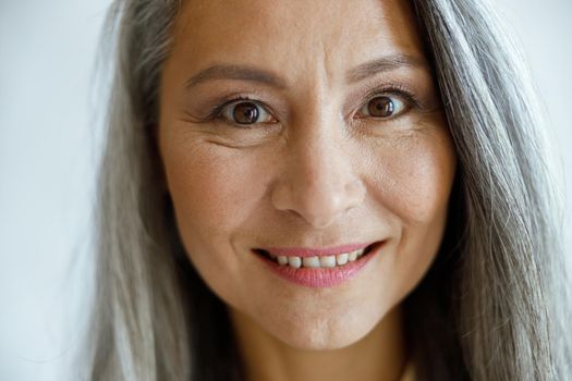 Cheerful silver haired lady with makeup looks into camera on light grey background