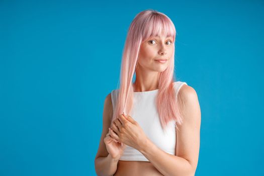 Young woman touching her smooth natural long pink dyed hair and looking surprised, posing isolated over blue studio background