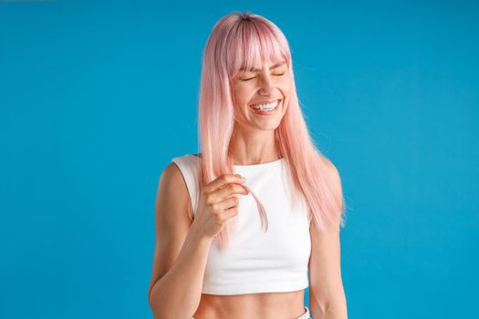 Joyful young woman with natural long pink dyed hair laughing with eyes closed, posing isolated over blue studio background