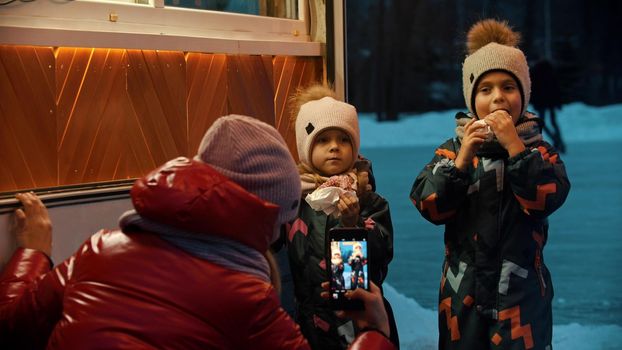A young woman taking photos of her children drinking hot drinks and eating donuts outdoors in winter