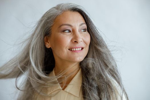 Happy middle aged Asian woman with flying grey locks stands on light background