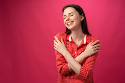 Joyful smiling young woman hugging herself against pink background