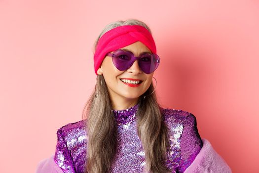 Fashion. Close up of stylish senior woman smiling happy at camera, wearing sunglasses and disco headband, pink background