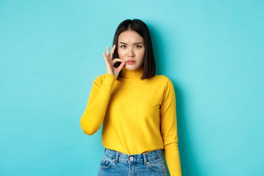 Serious asian girl showing mouth zip gesture, promise to keep shut and frowning, telling big secret, standing in yellow pullover over blue background