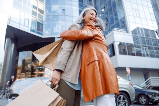Cheerful Asian woman hugs mature lady with numerous shopping bags on city street