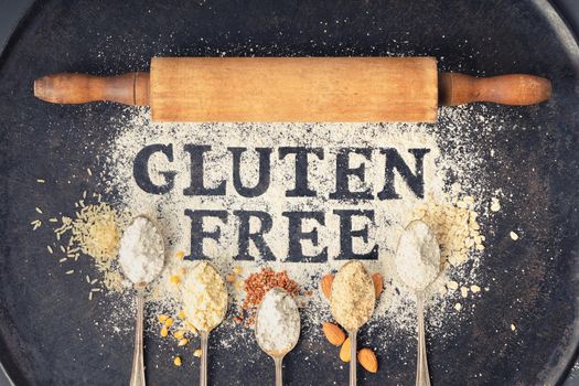 Gluten free written in flour on vintage baking sheet, rolling pin and spoons of various gluten free flour (almond flour, buckwheat flour, rice flour, corn flour, oatmeal flour), flat lay, top view