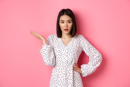 Careless asian woman raising hand and shrugging, staring at camera unbothered and questioned, so what gesture, standing over pink background