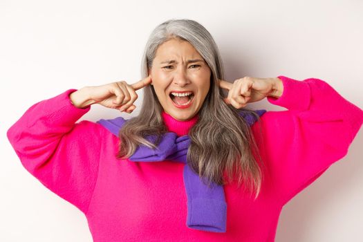 Close-up of annoyed and bothered asian senior woman screaming with shut ears, disturbed by loud noise, standing over white background