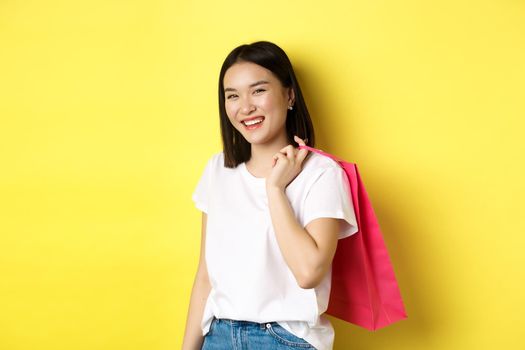 Happy asian woman holding shopping bag behind shoulder and laughing, standing over yellow background