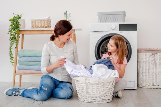 Mother with little daughter sorting clothes after laundry while sitting on floor in front of washing machine