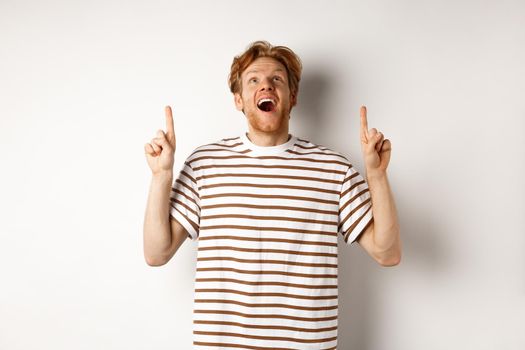 Fascinated redhead man looking with amazement and happiness, pointing fingers up, checking out something cool, standing over white background