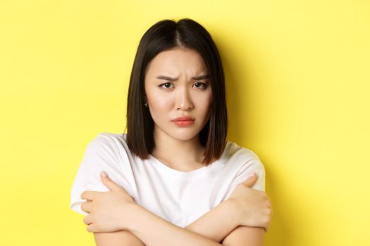 Sad timid girl comforting herself, hugging body and frowning upset, standing again yellow background