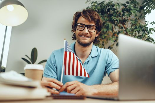 Young bearded businessman sitting at table with laptop and american flag