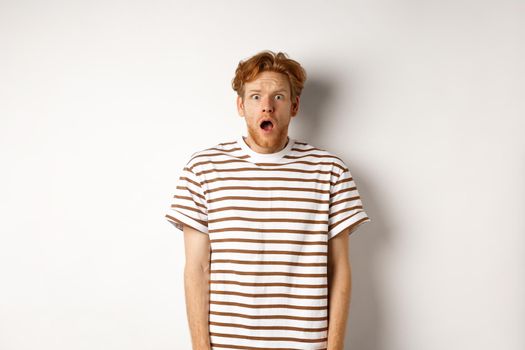 Shocked and scared redhead man screaming, open mouth and staring terrified at camera, standing over white background