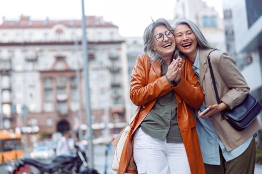 Joyful senior woman and Asian companion holding smartphone stand on modern city street