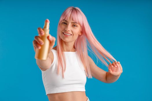 Portrait of young woman with pink hair smiling and showing bottle of moisturizing spray to camera while standing isolated over blue studio background