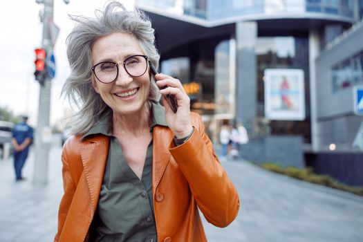 Happy grey haired senior woman with glasses talks on mobile phone on city street