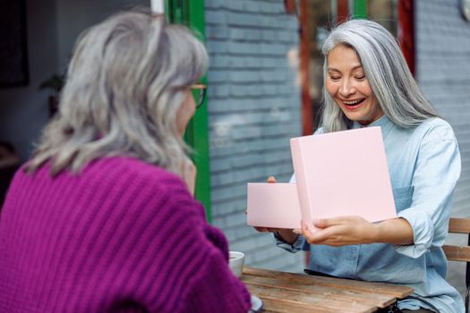 Happy senior Asian lady opens gift with best friend celebrating holiday in cafe