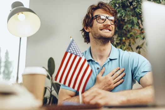 Young bearded businessman sitting at table with laptop and american flag