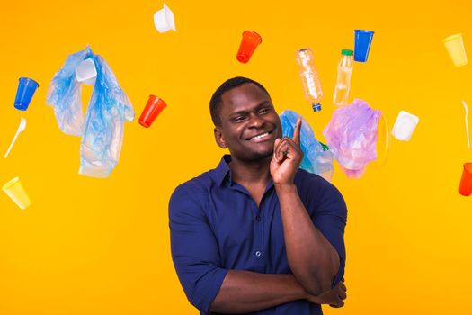 Problem of trash, plastic recycling, pollution and environmental concept - Serious african american man looking on trash on yellow background. He is thinking about ecology