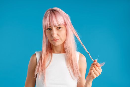 Young woman with natural long pink dyed hair holding a strand of it and looking upset at camera, posing isolated over blue studio background
