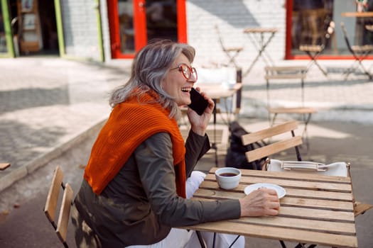 Positive grey haired senior lady talks on phone at table on outdoors cafe terrace