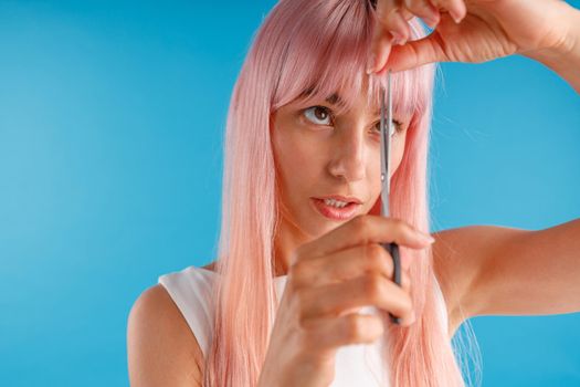 Portrait of cute young woman with pink hair cutting fringe herself using scissors, standing isolated over blue studio background