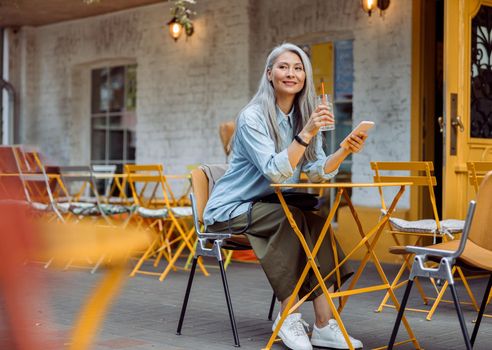 Grey haired Asian woman with glass and smartphone on outdoors cafe terrace