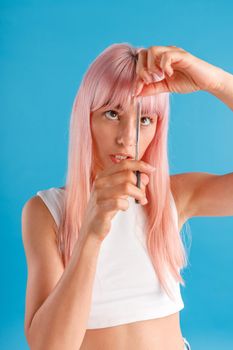 Cute young woman with pink hair cutting fringe herself using scissors, standing isolated over blue studio background