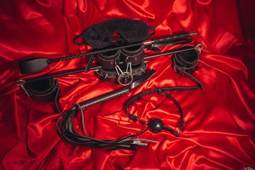 Bondage, kinky adult sex games, kink and BDSM lifestyle concept with a pair of leather handcuffs, flogger, ball gag and a coller with a leash attached on red silk with copy space