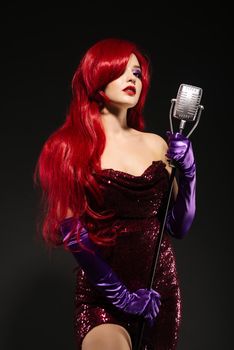 Young redhead woman with very long hair in red gown with microphone on the stand on a black background.