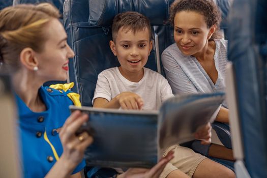 Female flight attendant entertaining a kid on board by offering a book to read. Cabin crew provide service to family in airplane