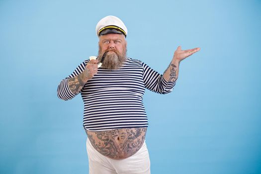 Dashing fat man with tattoos in sailor suit points aside on light blue background