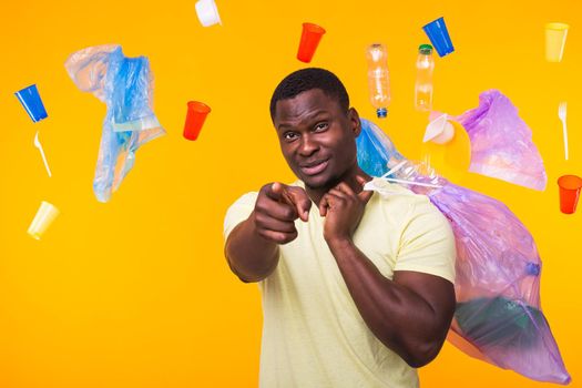 Problem of trash, plastic recycling, pollution and environmental concept - funny african american man carrying garbage for recycling and pointing on you on yellow background.