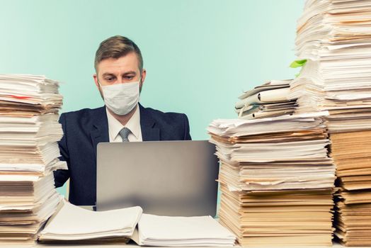 A male accountant or company manager works in an office in a pandemic in view of the accumulated paper work. A protective medical mask is on the face. On the desktop are large stacks of documents.