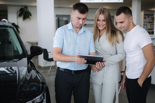Salesperson talking with couple and help choosing new car. Man and woman in car showroom. Man using tablet to shop opportunity of different models