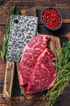 Raw denver cut black angus beef steak on a butcher board with meat cleaver. Dark wooden background. Top view