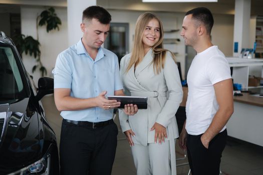 Salesperson talking with couple and help choosing new car. Man and woman in car showroom. Man using tablet to shop opportunity of different models