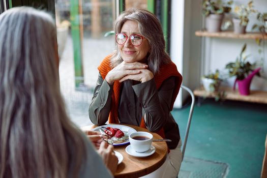 Grey haired woman with glasses looks at friend sitting at small table in cozy cafe