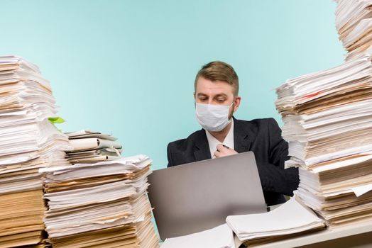 A male accountant or company manager works in an office in a pandemic in view of the accumulated paper work. A protective medical mask is on the face. On the desktop are large stacks of documents.