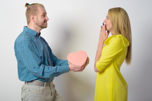 A stylish man in a checkered green shirt gives a beautiful slender girl in a yellow dress a gift box in the form of a heart.