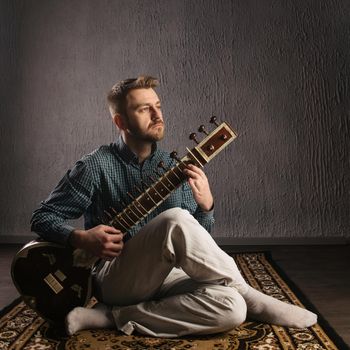 Portrait of a European man playing the sitar sitting on the carpet
