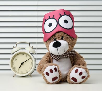 brown teddy bear sits with a bandage for sleeping on a white table. Near the alarm clock, time is seven in the morning, difficulty getting up in the morning