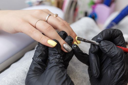 Nail master wearing black gloves applying brush on acrylic nails in the salon. Closeup on manicure
