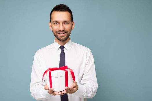 Shot of handsome positive smiling brunette unshaven young busibessman isolated over blue background wall wearing white shirt holding white gift box with red ribbon and looking at camera