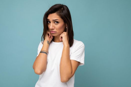 Portrait of sorrowful offended sad young pretty nice brunette woman with sincere emotions wearing casual white t-shirt for mockup isolated on blue background with copy space and covering ears with fingers