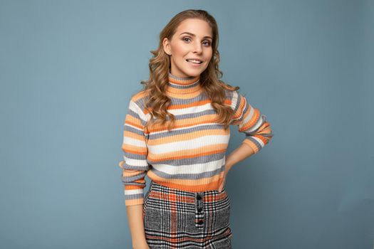 Young charming fascinating self-confident positive happy smiling blonde female person wearing casual striped pullover isolated over blue background with free space