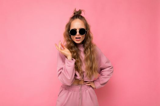 Fascinating dissatisfied adult blonde curly woman isolated over pink background wall wearing casual pink sport clothes and stylish sunglasses looking at camera