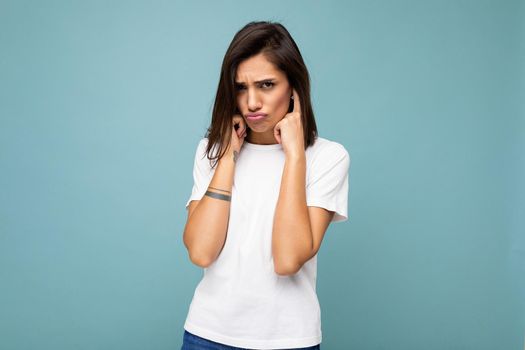 Portrait of touchu sad sorrowful young beautiful brunette woman with sincere emotions wearing casual white t-shirt for mockup isolated on blue background with empty space and covering ears
