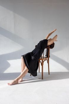 young girl in black dress poses on chair on white cyclorama in pattern of light and shadow near window bending her body
